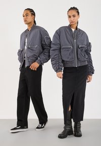 Twee modellen met een vlecht in hun haar, gekleed in grijze bomberjacks; de één in zwarte broeken en sneakers, de ander in een zwarte rok en laarzen.