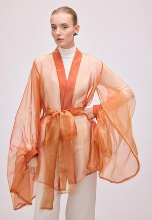 Le modèle pose dans un kimono vaporeux orange pêche, à manches amples et ceinturé d’une ceinture nouée, porté sur un haut et un pantalon blancs.
