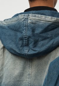 Μπουφάν από τζιν με κουκούλα, σε δίχρωμο σχέδιο με σκούρο και ανοιχτό μπλε, χαρακτηρίζεται από υφή υφάσματος και εμφανείς ραφές.