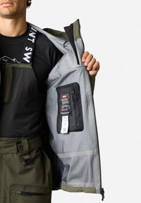 Wasserfeste Jacke in Olivgrün mit einer Reißverschlusstasche im Inneren. Verfügt über ein glattes graues Futter und ein sichtbares Etikett neben einem schwarzen Hardware-Akzent.