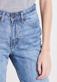 Jeans de mezclilla azul claro con cintura alta, que cuentan con cremallera, cierre de botón metálico y un desvanecimiento sutil. Acentos de costura notables.