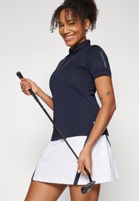 Tumed polo särk krae ja lühikeste varrukatega, koos valge seelikuga. Omab logo varrukal ja golfikeppi käes.