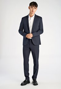 Granatowy garnitur z dopasowaną marynarką i zwężanymi spodniami, w połączeniu z białą koszulą. Całość uzupełniają czarne eleganckie buty. Gładka tekstura tkaniny.