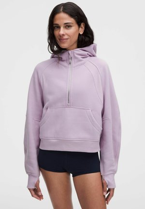 Kvinde iført en lavendel halv-zip hættetrøje med kængurulomme og sorte shorts, stående mod en ensfarvet lys baggrund.