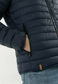Dunkelblaue Puffjacke mit gestepptem Muster, Reißverschluss-Taschen und einem braunen Lederakzent am Saum. Entwickelt für Isolierung und Wärme.