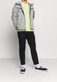 Man som bär en ljusgrå zip-up jacka, neon gul t-shirt, svarta byxor, vita strumpor och svarta sneakers som står framför en helt vit bakgrund.