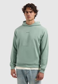 Man in een mintgroene hoodie met de tekst "PURE PATH", lichte gedistresseerde jeans, staand tegen een effen lichtgrijze achtergrond.