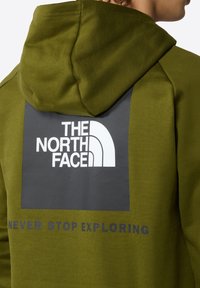 Felpa con cappuccio verde oliva caratterizzata da un grande rettangolo grigio con il logo "THE NORTH FACE" e il testo "NEVER STOP EXPLORING" in grigio scuro sottostante.