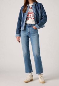 Veste en jean avec manches plus claires, t-shirt blanc à motif rouge, jean bleu clair droit et baskets beiges.