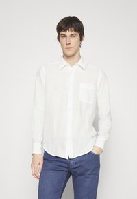 Camisa blanca de lino de manga larga con botones en la parte delantera, un bolsillo en el pecho y un dobladillo ligeramente curvado, combinada con pantalones de mezclilla azules.