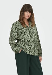 Blusa verde a fantasia con una vestibilità comoda, maniche lunghe, colletto e macchie astratte nere. Abbinata a pantaloni verde scuro.