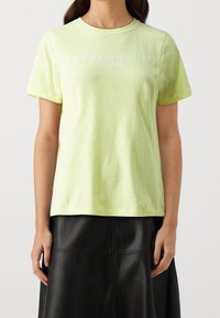 Mujer vistiendo una camiseta ligera amarilla de Tommy Hilfiger con texto de logo y una falda de cuero negra, de pie contra un fondo sencillo.