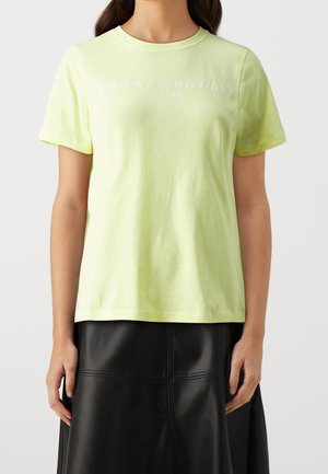 Woman wearing a light yellow Tommy Hilfiger t-shirt with logo text and a black leather skirt, standing against a plain background.