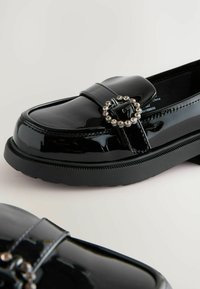 Mocasini din piele patent neagră cu o aplicație rotundă din argint, talpă din cauciuc plat și finisaj neted; design clasic cu braț decorativ.