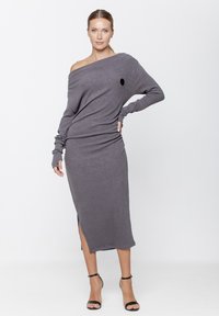 Szara, prążkowana sukienka dzianinowa z dekoltem na jedno ramię, długimi rękawami z otworami na kciuki oraz bocznym rozcięciem. Modelka ma na sobie czarne sandały na cienkich paskach.
