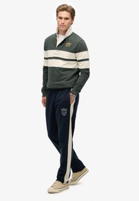 Groene en crèmekleurige gestreepte pullover met een kraag, gecombineerd met marineblauwe joggers met zijstrepen en logo. Tan sneakers maken de outfit af.