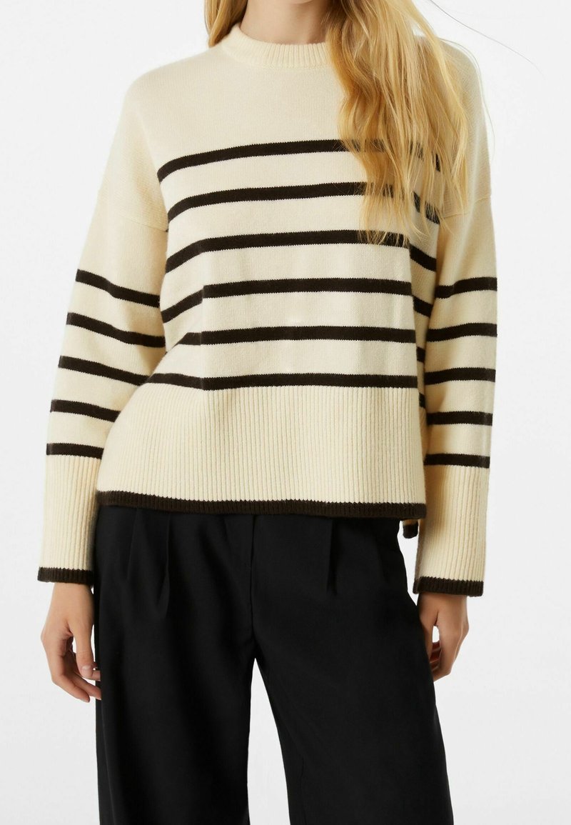 Femme portant un pull crème avec des rayures horizontales marron foncé et des poignets côtelés, associé à un pantalon noir ample.