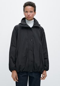 Giacca nera a maniche lunghe realizzata in tessuto leggero, con collo alto, chiusura a bottoni ed orlo elasticizzato, indossata sopra un dolcevita bianco.