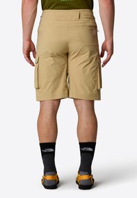 Pantaloni cargo beige realizzati in materiale leggero con tasche laterali. Presentano una vestibilità dritta e dettagli cuciti. Indossati con calze nere e sandali.