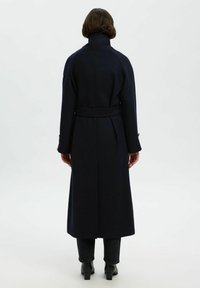 Cappotto lungo in lana blu navy con un colletto alto, vita cinturata e tasche laterali. Presenta una linea dritta con uno spacco sul retro per facilitare i movimenti.