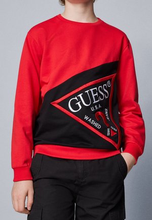 Barn iført en rød sweatshirt med en stor sort trekantet GUESS-logo og sorte bukser, stående med den ene hånd i lommen.