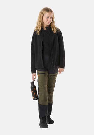 Junge Person mit lockigem blondem Haar, die eine schwarze Jacke, grüne und schwarze Outdoor-Hose trägt und eine schwarze Wasserflasche mit orangefarbenen Elch-Motiven hält.