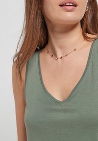 Débardeur ajusté vert olive avec un décolleté en V, doté d'une texture lisse. Collier en or avec de petites accents en forme d'étoile.