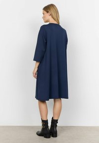 Marineblauwe jurk van jersey met driekwartmouwen en een ontspannen pasvorm, voorzien van een ronde halslijn en een gladde textuur. Zwarte enkellaarsjes met gespen.
