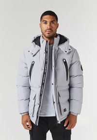 Light grey puffer jacket with a hood, featuring black zippers, front pockets, and branding on the zipper tape. Made from a smooth material.