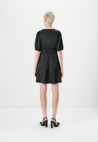 Vestido preto curto com mangas bufantes, cintura elástica e uma textura suave. O vestido cai acima do joelho, combinado com sapatos de salto pretos.