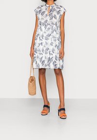 Witte jurk met blauw bladerenpatroon, keyhole-halslijn en korte mouwen. Gecombineerd met oranje en marineblauwe hoge hakken en een gevlochten tas.