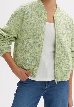 Frau trägt eine hellgrüne, strukturierte Zip-up-Jacke über einem weißen, gerippten Tanktop und blauen Jeans, mit einer Hand in der Tasche.