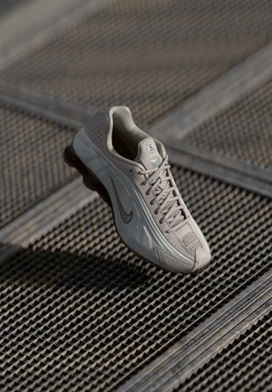 Lysegrå Nike-sneaker med sort swoosh-logo hængende over et metalristgulv, der viser side- og topdetaljer.