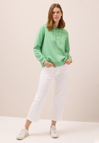 Felpa verde con cappuccio e lacci, abbinata a pantaloni bianchi tagliati e sneakers beige. Design semplice, texture morbida e vestibilità rilassata.