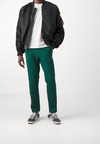 Černá bomber bunda na zip, zelené kalhoty s bočními pruhy a šedé tenisky. Pod bundou bílý tričko.