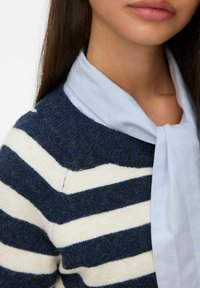 Pull rayé bleu marine et blanc avec un tricot texturé, associé à une chemise à col bleu clair nouée au cou. Vue en gros plan de l'encolure.
