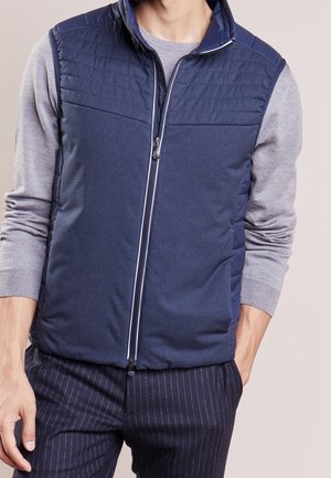 Man draagt een marineblauw doorgestikt gilet met rits over een lichtgrijs shirt met lange mouwen en donkere krijtstreep broek, met één hand in de zak.