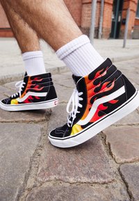 Høje sneakers med et sort ruskinds overdel, orange og rødt flamme design, hvide snørebånd og en hvid ydersål, sat mod en brostensbelagt overflade.