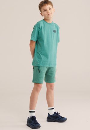 Fiatal fiú áll hátrahajtott kézzel, színeszöld pólót és rövidnadrágot visel, fehér zoknival és sötétkék tornacipővel, egyszínű háttér előtt.