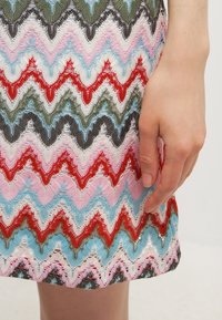 Robe tricotée arborant un motif en zigzag aux couleurs rouge, bleu, vert, rose et blanc, avec une apparence texturée semblable à de la dentelle. Main placée sur le tissu.