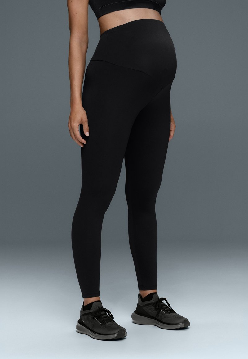 Femme enceinte portant des leggings de maternité noirs et des chaussures de sport noires, debout sur un fond dégradé gris.