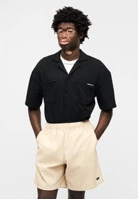 Μαύρο πουκάμισο με κοντά μανίκια και υφή υφάσματος, με δύο τσέπες μπροστά, συνδυασμένο με μπεζ ελαστικό σορτς. Και τα δύο ρούχα είναι απλά και casual.