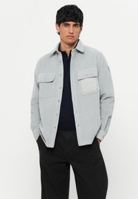 Light gray corduroy shirt jacket with two chest pockets, button closure, and a plain texture, paired with a navy inner garment and black pants.