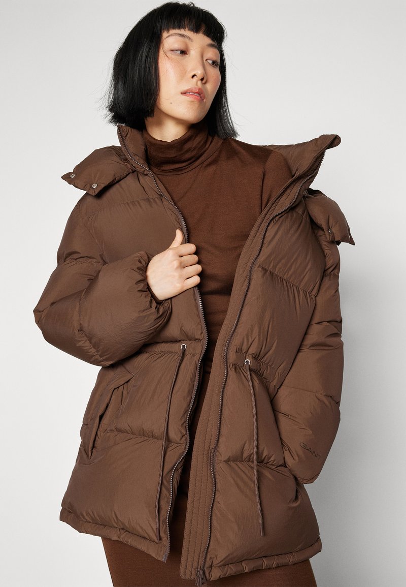 Brązowa kurtka puchowa z wysokim kołnierzem, podwójnym kapturem oraz sznurkami. Posiada duże pikowane sekcje i boczne kieszenie.