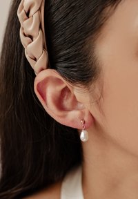 Perlenohrringe mit einer sphärischen goldenen Oberseite und einer hängenden ovalen weißen Perle; getragen mit einem geflochtenen beigen Stoffkopfband.