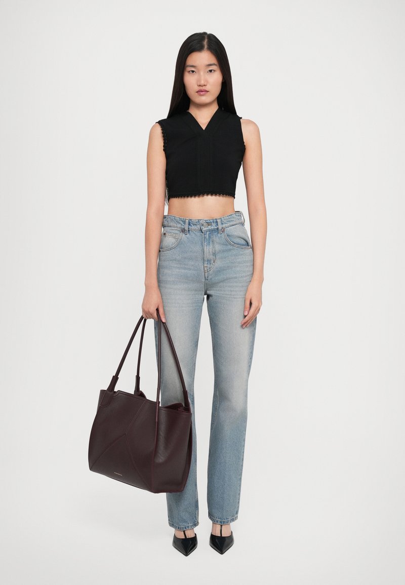 Femme portant un crop top sans manches noir, un jean taille haute bleu clair, des escarpins noirs pointus, tenant un grand sac fourre-tout en cuir brun foncé.