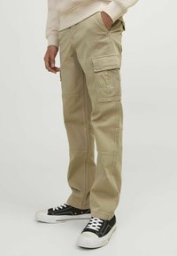 Beige cargo byxor med flera fickor, sydda detaljer, raka ben och ett hållbart bomullsblandningsmaterial. Kombineras med svarta sneakers.