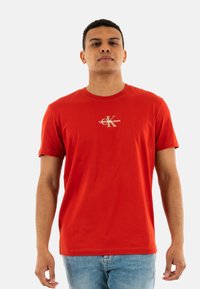 Camiseta de algodón roja con cuello redondo y mangas cortas. Presenta un pequeño logotipo de "Calvin Klein Jeans" en blanco y dorado en el pecho.