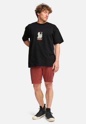 Svart kortermet T-skjorte med et sentralt grafisk design i fargene teal, oransje og hvit, sammen med rustfargede shorts og svarte joggesko.