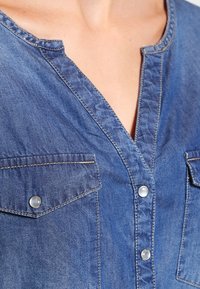 Chemise en denim avec un col en V, deux poches poitrine à fermeture pression et des coutures contrastantes en bleu clair. Texture douce avec une coupe décontractée.